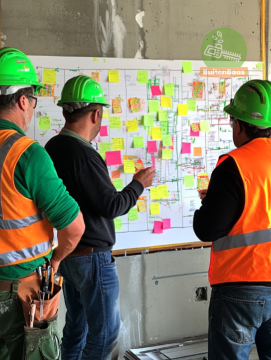 Drie mannen in oranje hesjes met een groene bouwhelm op, die met elkaar de planning bespreken. Ze staan voor een groot bord waar allemaal post-it's op geplakt zijn.